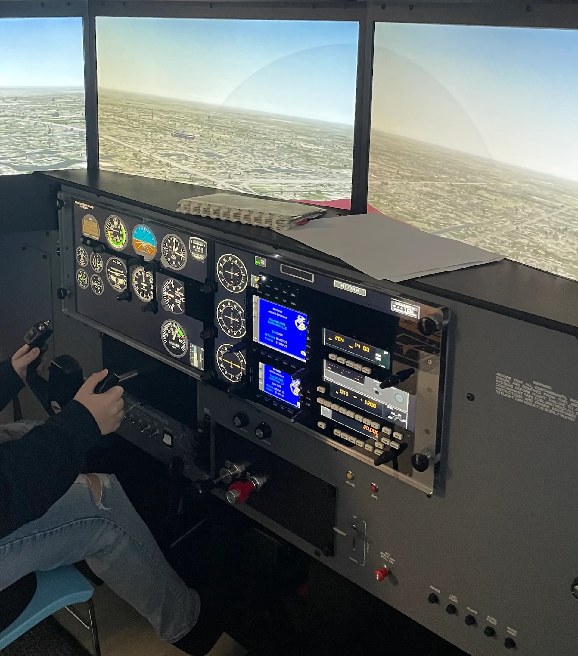 Hands on the controls of a Redbird flight simulator in front of a Cessna-style instrument panel and three screens at Sun City Aviation Academy